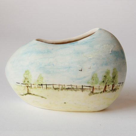 Ceramic vase with a painted landscape of trees and a fence against a blue sky.